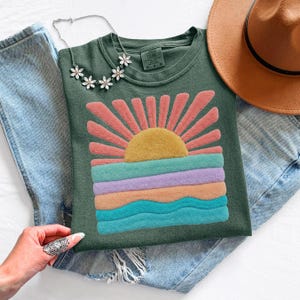 May include: A dark green t-shirt featuring a textured, raised graphic of a sun rising over a multi-colored landscape of horizontal stripes in yellow, pink, teal, lavender, and orange. The shirt is paired with blue denim jeans and a brown hat.