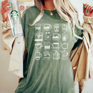 May include: A green t-shirt featuring a grid of white line drawings of coffee-related items, including cups, pots, and machines. The shirt has a relaxed fit and is made of a soft material. A clear Starbucks tumbler is held in the left hand.