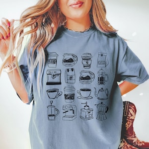 May include: A light blue t-shirt featuring a collection of hand-drawn coffee-related illustrations. The design includes various coffee cups, coffee makers, and coffee pots. The shirt is a comfortable, casual style.