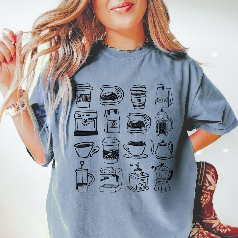 May include: A light blue t-shirt featuring a collection of hand-drawn coffee-related illustrations. The design includes various coffee cups, coffee makers, and coffee pots. The shirt is a comfortable, casual style.