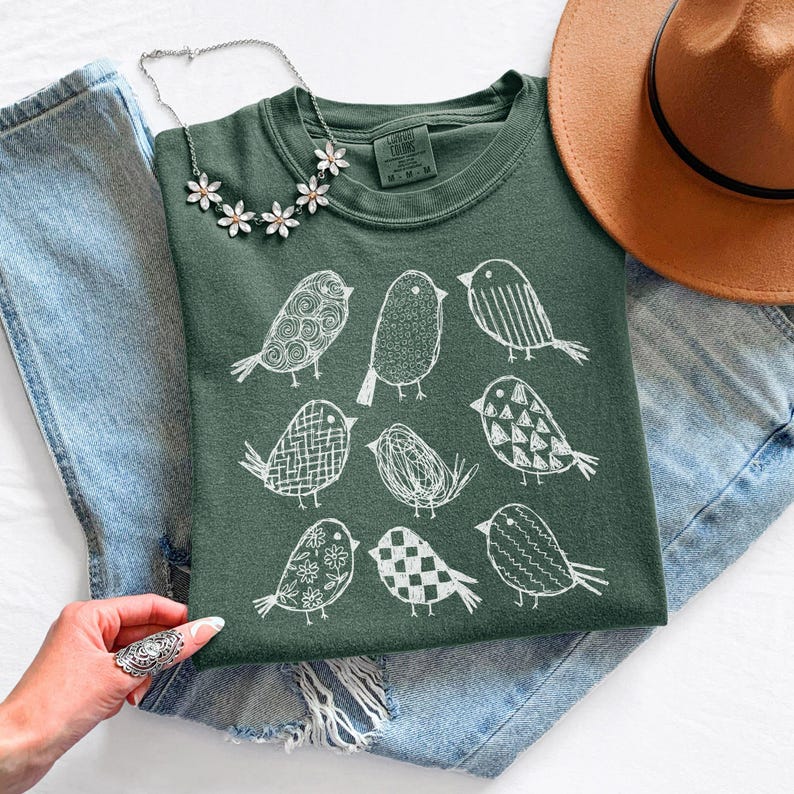 May include: A dark green t-shirt featuring a white illustration of nine birds with various patterns. The shirt is folded on top of blue denim jeans, with a brown hat and a floral necklace visible.
