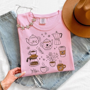 Peut inclure: T-shirt rose clair Comfort Colors avec des illustrations sur le th&egrave;me du caf&eacute;, comprenant des th&eacute;i&egrave;res, des tasses &agrave; caf&eacute; et une cafeti&egrave;re. Le design comprend &eacute;galement des motifs floraux. Un chapeau marron et un collier argent&eacute; sont &eacute;galement visibles.