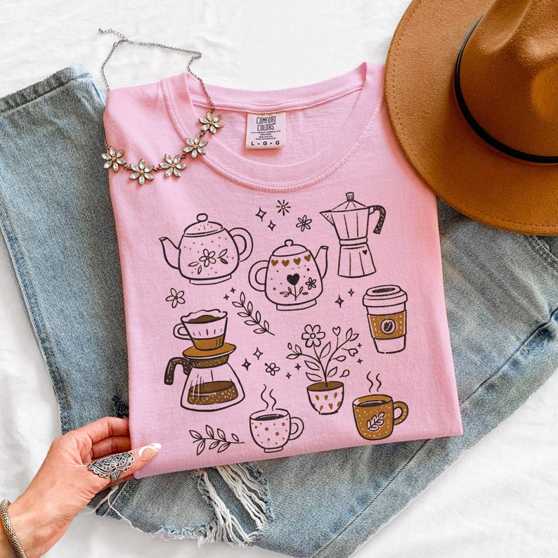 Peut inclure: T-shirt rose clair Comfort Colors avec des illustrations sur le th&egrave;me du caf&eacute;, comprenant des th&eacute;i&egrave;res, des tasses &agrave; caf&eacute; et une cafeti&egrave;re. Le design comprend &eacute;galement des motifs floraux. Un chapeau marron et un collier argent&eacute; sont &eacute;galement visibles.