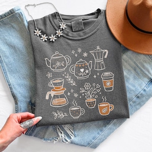 Peut inclure: Sweat-shirt gris fonc&eacute; avec des illustrations blanches et orange de th&eacute;i&egrave;res, cafeti&egrave;res, tasses &agrave; caf&eacute; et plantes. Un collier argent&eacute; &agrave; fleurs et un chapeau marron sont &eacute;galement visibles.