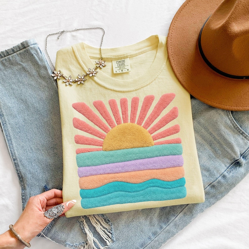 May include: A pale yellow t-shirt featuring a textured, fuzzy graphic of a sun rising over ocean waves in shades of pink, yellow, lavender, peach, and teal. The shirt has a crew neckline and a Comfort Colors label. Also includes blue jeans, a brown hat, and a silver-toned floral necklace.