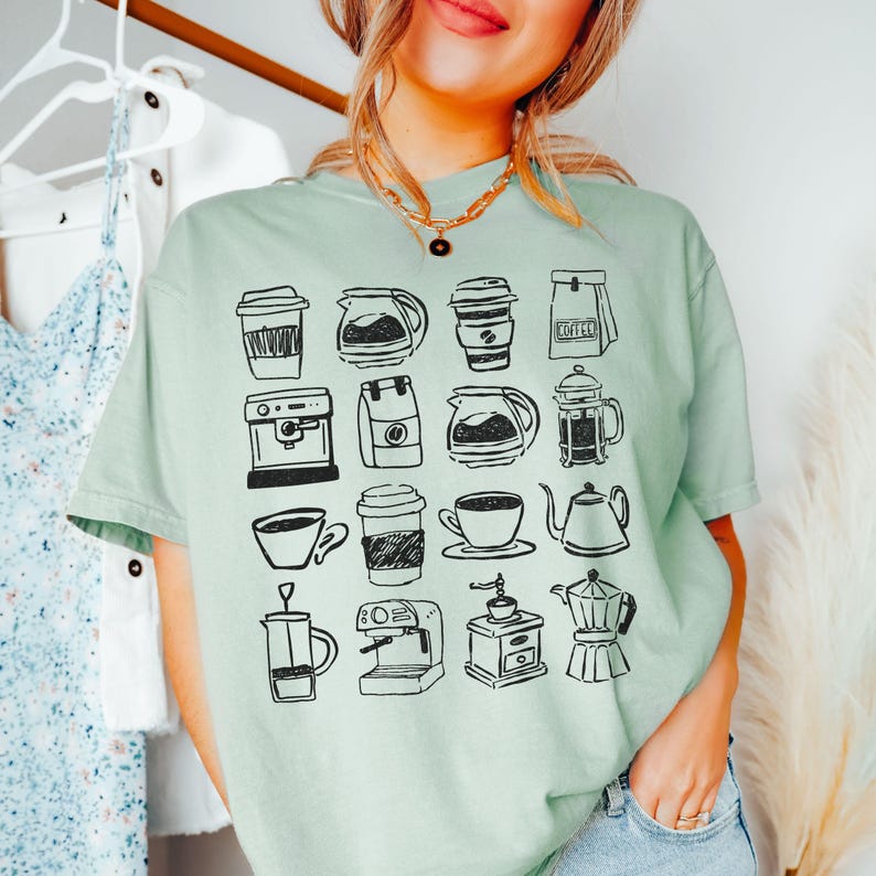 May include: A light green t-shirt featuring a black and white illustration of various coffee-related items. The design includes coffee cups, coffee makers, coffee pots, and coffee bean bags. The shirt is a comfortable, casual style.