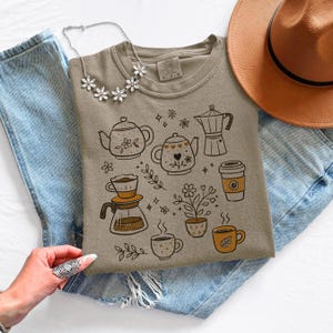 Peut inclure: Un sweat-shirt col rond de couleur beige avec un motif sur le th&egrave;me du caf&eacute; et du th&eacute;. Le motif comprend des illustrations de th&eacute;i&egrave;res, de cafeti&egrave;res, de tasses et d'&eacute;l&eacute;ments floraux. Le sweat-shirt est pr&eacute;sent&eacute; sur un jean bleu.