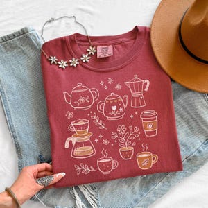 Peut inclure: Un t-shirt bordeaux avec un motif blanc repr&eacute;sentant des &eacute;l&eacute;ments li&eacute;s au caf&eacute; et au th&eacute;, comme des th&eacute;i&egrave;res et des tasses &agrave; caf&eacute;. Le t-shirt est associ&eacute; &agrave; un chapeau marron, un collier floral et un jean bleu. L'&eacute;tiquette indique "Comfort Colors XL TG-EG".
