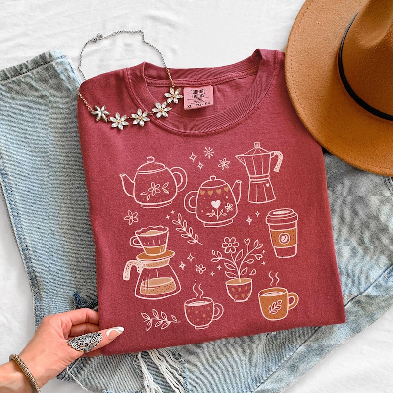 Peut inclure: Un t-shirt bordeaux avec un motif blanc repr&eacute;sentant des &eacute;l&eacute;ments li&eacute;s au caf&eacute; et au th&eacute;, comme des th&eacute;i&egrave;res et des tasses &agrave; caf&eacute;. Le t-shirt est associ&eacute; &agrave; un chapeau marron, un collier floral et un jean bleu. L'&eacute;tiquette indique "Comfort Colors XL TG-EG".