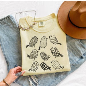 May include: A light yellow t-shirt featuring nine black bird illustrations with various patterns. The shirt is laid on top of blue jeans, with a brown hat and a floral necklace nearby. The shirt has the text "COMFORT COLORS" on the tag.