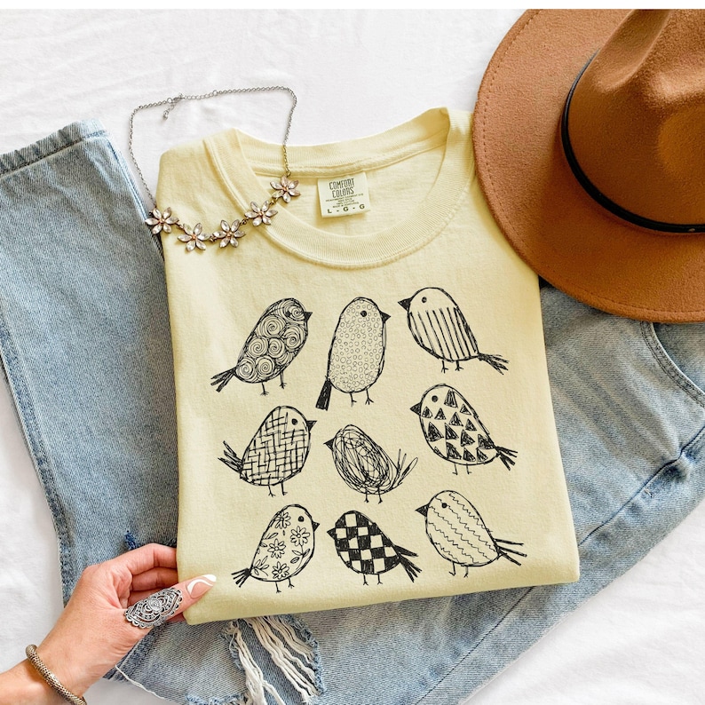 May include: A light yellow t-shirt featuring nine black bird illustrations with various patterns. The shirt is laid on top of blue jeans, with a brown hat and a floral necklace nearby. The shirt has the text "COMFORT COLORS" on the tag.