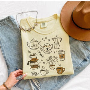 Peut inclure: Un t-shirt jaune clair Comfort Colors avec des illustrations sur le th&egrave;me du caf&eacute; en marron. Le motif comprend des th&eacute;i&egrave;res, des tasses &agrave; caf&eacute;, une cafeti&egrave;re et un gobelet &agrave; caf&eacute; &agrave; emporter. Le t-shirt est associ&eacute; &agrave; un jean bleu, un chapeau marron et un collier floral.