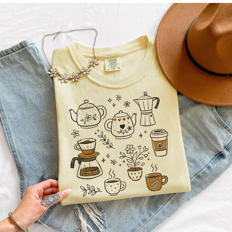 Peut inclure: Un t-shirt jaune clair Comfort Colors avec des illustrations sur le th&egrave;me du caf&eacute; en marron. Le motif comprend des th&eacute;i&egrave;res, des tasses &agrave; caf&eacute;, une cafeti&egrave;re et un gobelet &agrave; caf&eacute; &agrave; emporter. Le t-shirt est associ&eacute; &agrave; un jean bleu, un chapeau marron et un collier floral.