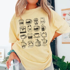 May include: A light yellow t-shirt featuring a black and white illustration of various coffee-related items, including coffee makers, cups, and coffee bags. The shirt is a relaxed fit.