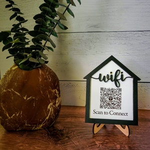 May include: A black wooden sign with a white background and the word "wifi" written in cursive. A QR code is displayed on the sign with the text "Scan to Connect" below it. The sign is on a wooden stand and is in front of a white wooden wall.