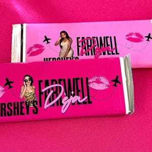 May include: Two Hershey's chocolate bars with pink wrappers. The wrappers feature the text "FAREWELL Deja" in a cursive font, along with images of a woman, a kiss, and an airplane. The background is a solid pink color.