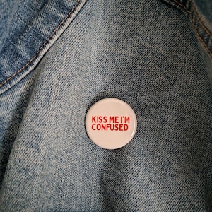 May include: A white button with the text "KISS ME I'M CONFUSED" in red lettering. The button is pinned to a blue denim jacket.
