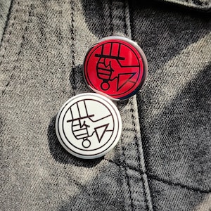 May include: Two round button pins with a black and red design. One pin is red with a black graphic, and the other is white with a black graphic. The pins are on a gray denim jacket.