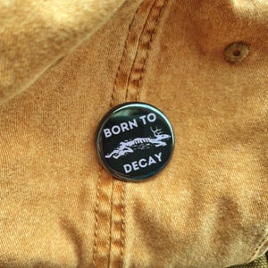May include: A black button with the text "BORN TO DECAY" in white, along with a white graphic of a deer skeleton. The button is set against a tan fabric background, likely a hat or similar item.