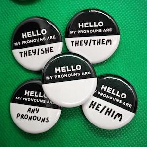 May include: A collection of black and white pronoun buttons. Each button has the text "HELLO MY PRONOUNS ARE" above a pronoun set, including "THEY/HE", "THEY/SHE", "THEY/THEM", "ANY PRONOUNS", and "HE/HIM".