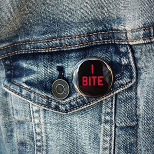 May include: A black button pin with the text "I BITE" in red letters is pinned to a blue denim jacket pocket. The button has a glossy finish and is round. The jacket has a button closure and visible stitching.