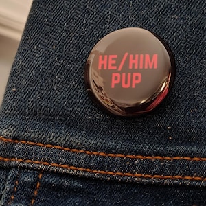 May include: A round, dark brown button with the text "HE/HIM PUP" in red. The button is pinned to a blue denim jacket. The button has a glossy finish and reflects the surrounding environment.
