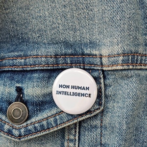 May include: A white button pinned to a denim jacket. The button has the text "NON HUMAN INTELLIGENCE" in black, centered on the button. The jacket is blue with visible stitching and a metal button.