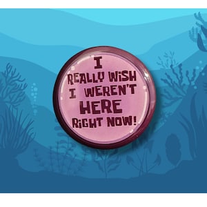 May include: A round, pink button with the text "I REALLY WISH I WEREN'T HERE RIGHT NOW!" in a bold, black font. The button has a dark pink border and is set against a blue background with aquatic plant silhouettes.