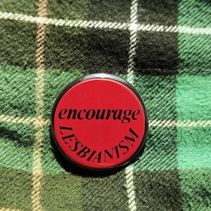 May include: A round, red button with the words "encourage LESBIANISM" in black text. The button is set against a green and black plaid background.