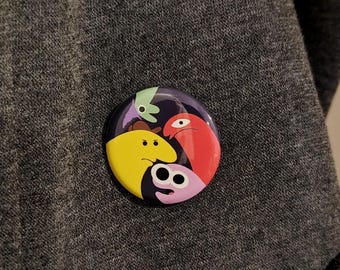 May include: A round button pin featuring a colorful cartoon design. The pin has a dark purple background with four cartoon characters in yellow, red, pink, and green. The pin is resting on a gray fabric surface.