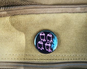 May include: A round, black button with a glossy finish. The button features four cartoon ghost figures with glasses, outlined in pink. The button is set against a textured, tan fabric background with a zipper.