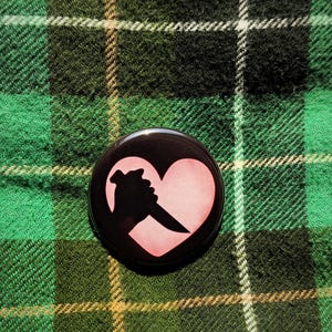 May include: A black button with a graphic of a pink heart with a knife silhouette. The button is set against a green and black plaid background.