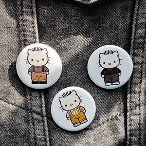 May include: Three white round buttons featuring a cartoon cat character in different outfits. The cat is wearing brown overalls, a black shirt, and a yellow and white striped shirt with brown overalls. The buttons are on a gray denim jacket.