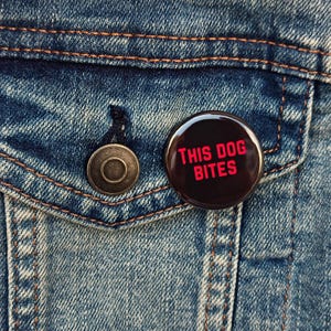 May include: A round, black button with the text "THIS DOG BITES" in red lettering. The button is pinned to a blue denim jacket, near a metal button and a pocket.
