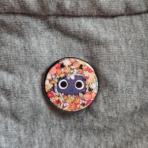 May include: A round button pin featuring a cartoon character with large eyes and a floral wreath design. The pin has a black border and is set against a gray fabric background. The flowers are in shades of red, orange, yellow, and white.