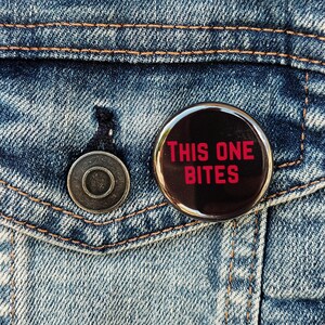 May include: A round, black button with the text "THIS ONE BITES" in red letters. The button is pinned to a blue denim jacket. The jacket has a metal button and orange stitching.
