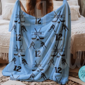 May include: A light blue fleece blanket with a hockey theme. The blanket features a repeated pattern of hockey sticks, pucks, and the number 12. The name "Justin" is printed on the blanket.