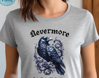 May include: A light gray t-shirt featuring a detailed illustration of a dark blue raven with the text "Nevermore" above and "But Make It Fashion" below. The raven is surrounded by ornate floral designs. The shirt has short sleeves.