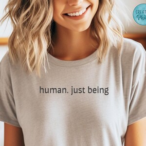 May include: A light gray t-shirt with the words "human. just being" printed in black. The shirt is short-sleeved and appears to be made of a soft material. A circular logo with the text "Creative Philly" is visible in the upper right corner.