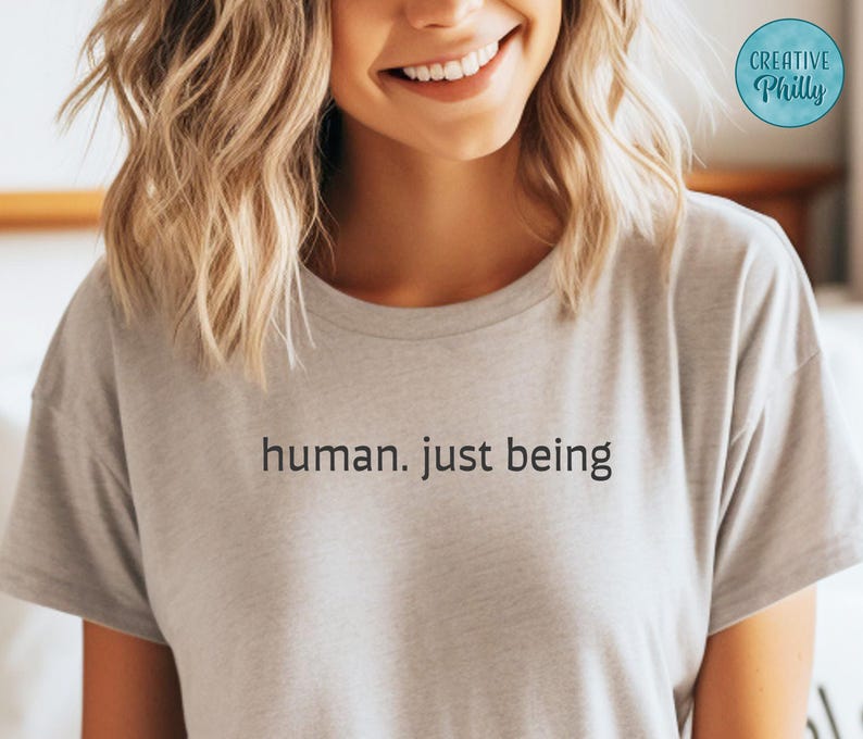 May include: A light gray t-shirt with the words "human. just being" printed in black. The shirt is short-sleeved and appears to be made of a soft material. A circular logo with the text "Creative Philly" is visible in the upper right corner.