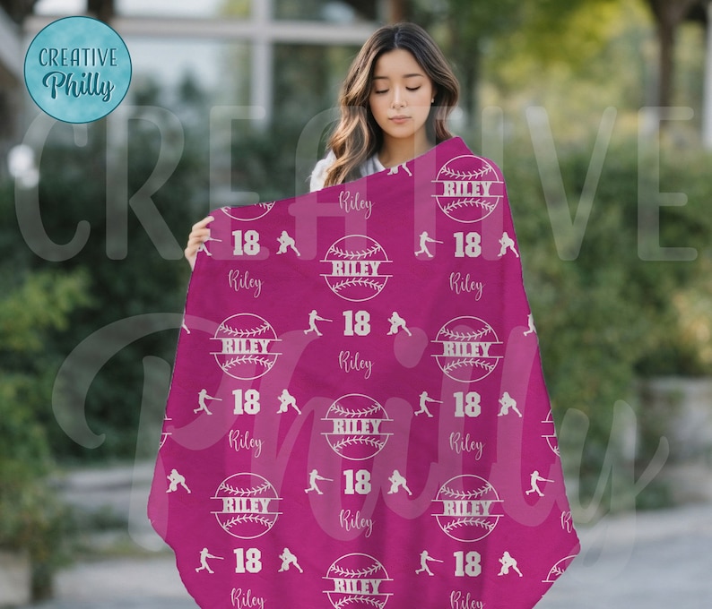 May include: A pink fleece blanket with a white baseball design. The blanket has the name Riley printed on it, along with the number 18. The blanket is folded in half and is being held by a person.