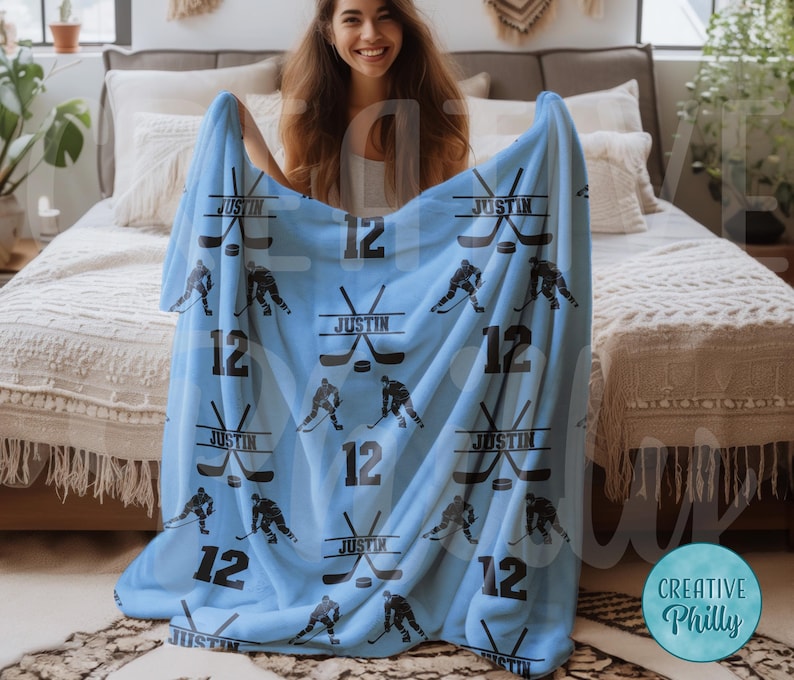 May include: A light blue fleece blanket with a hockey theme. The blanket features a repeated pattern of hockey sticks, pucks, and the number 12. The name "Justin" is printed on the blanket.