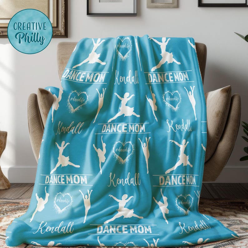 May include: A turquoise blue fleece blanket with a white silhouette of a dancer repeated across the fabric. The blanket has the words "Dance Mom" and "Kendall" printed in white on the blanket.