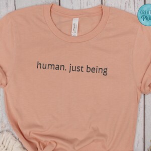 May include: A peach-colored t-shirt with the words "human. just being" printed in black. The shirt has a crew neck and short sleeves. The brand tag is visible on the upper left side of the shirt.