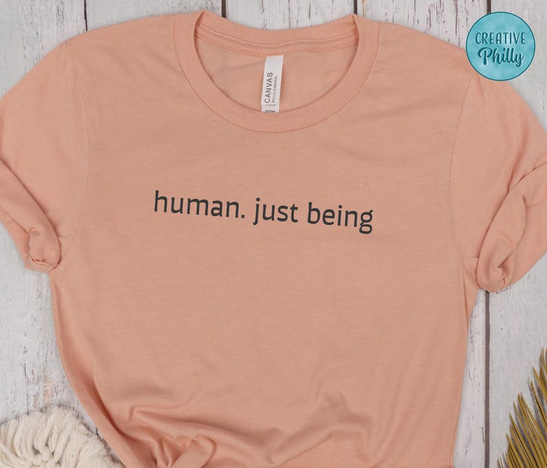 May include: A peach-colored t-shirt with the words "human. just being" printed in black. The shirt has a crew neck and short sleeves. The brand tag is visible on the upper left side of the shirt.