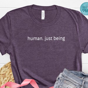 May include: A purple heather t-shirt with the text "human. just being" in white. The shirt has a crew neck and short sleeves. The shirt is made of a soft, comfortable material.