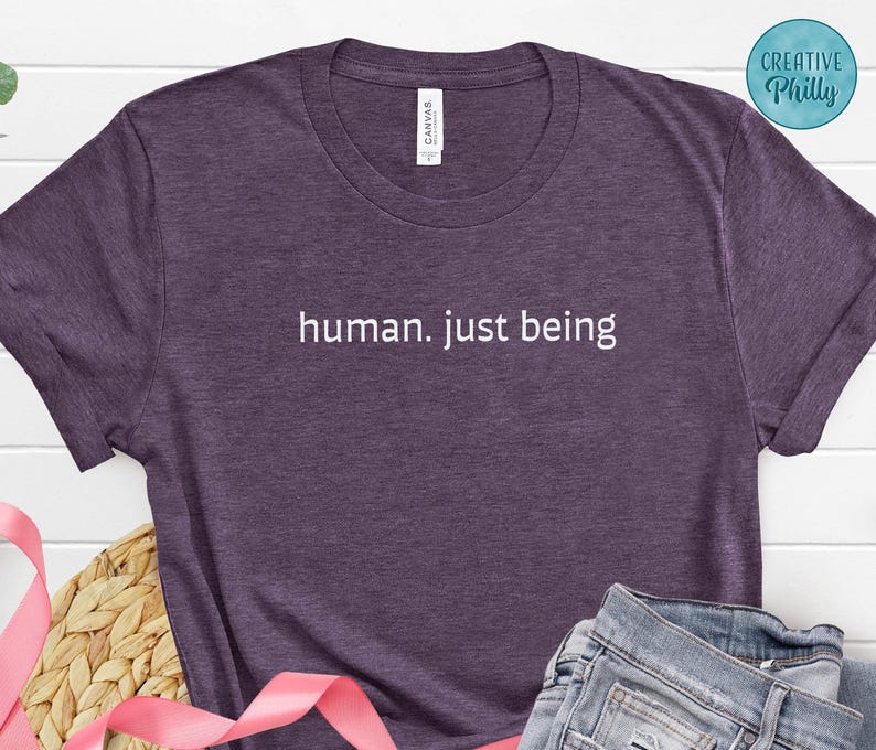 May include: A purple heather t-shirt with the text "human. just being" in white. The shirt has a crew neck and short sleeves. The shirt is made of a soft, comfortable material.