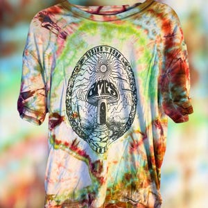 May include: A short-sleeve t-shirt with a multi-colored tie-dye pattern in shades of green, blue, orange, and brown. The center features a black graphic of a mushroom inside an oval frame with the text SPRING TOUR 2024 and the word OMES.