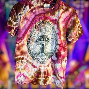 May include: A short-sleeve t-shirt with a tie-dye pattern in shades of burgundy, pink, and cream. The center features a black graphic print of a mushroom house, a stone path, and the text "SPRING TOUR" and "2026" inside an oval frame.