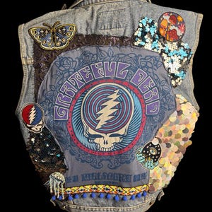 Puede incluir: Un chaleco vaquero con varios parches y adornos. La parte trasera presenta un gran gráfico de una calavera con un rayo, rodeado por las palabras "Grateful Dead". Parches adicionales incluyen una mariposa, setas y lentejuelas. El dobladillo inferior tiene pompones azules.